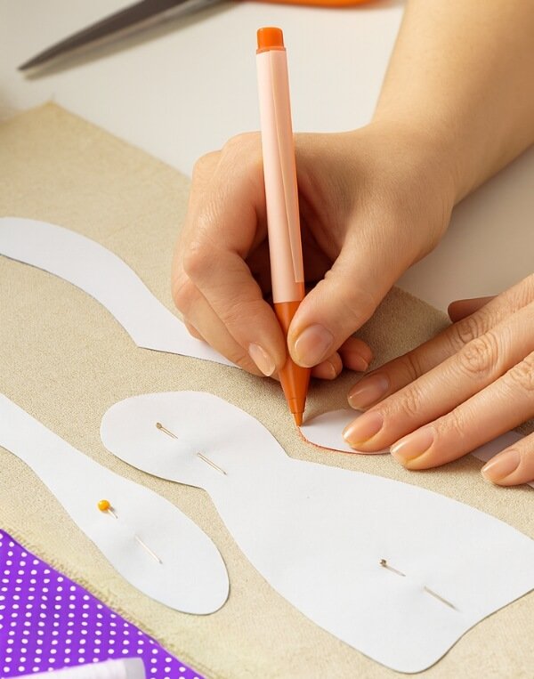 Mãos traçando molde de costura sobre tecido bege com caneta laranja, representando o processo técnico e detalhista de criação.
